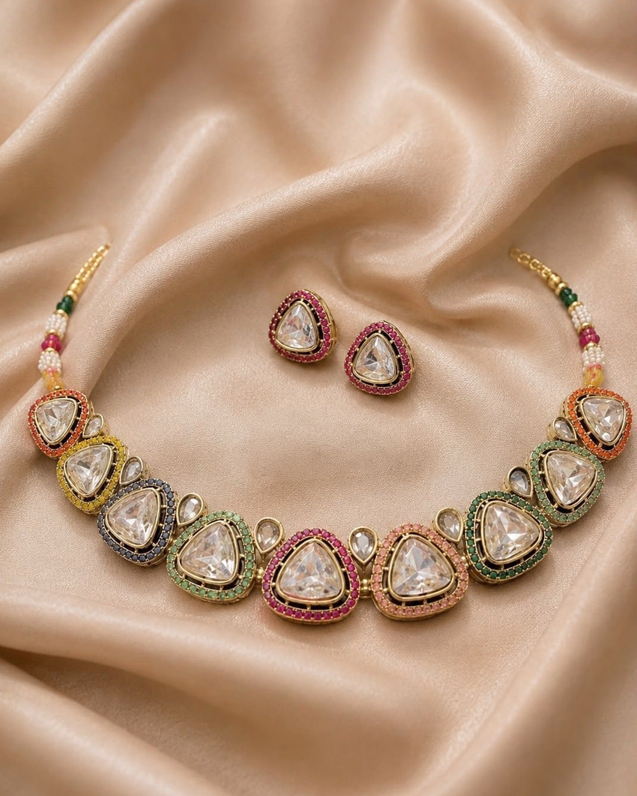 A traditional Indian Kundan necklace and matching stud earrings displayed on draped beige satin fabric. The necklace features clear teardrop-shaped stones set in gold with colorful accents of green, pink, orange, and blue, along with small white beads.