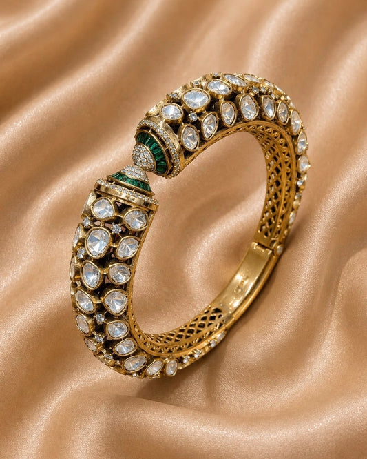 An ornate gold bangle bracelet adorned with clear stones and green emerald accents 