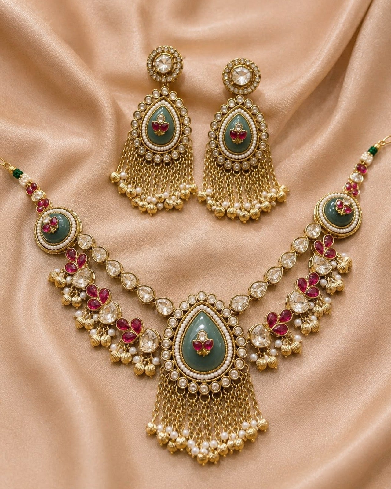 Gold-toned traditional Indian jewelry set including a necklace and matching earrings, decorated with green stones, red floral accents, and white pearl-like beads, displayed on a beige satin fabric background.