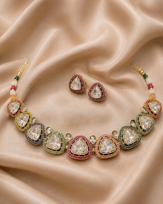 A traditional Indian Kundan necklace and matching stud earrings displayed on draped beige satin fabric. The necklace features clear teardrop-shaped stones set in gold with colorful accents of green, pink, orange, and blue, along with small white beads.