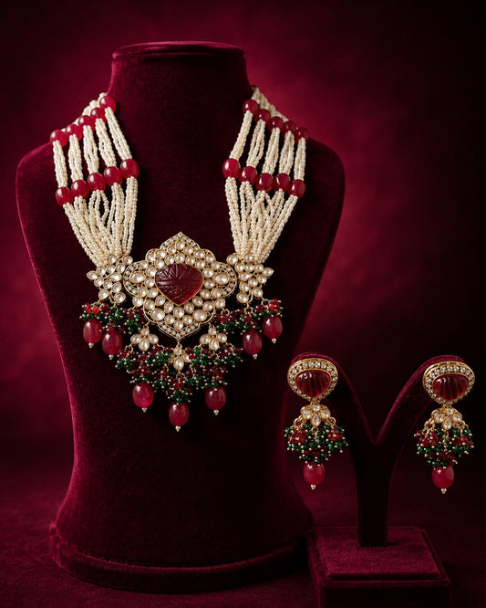Elegant ruby red and green beaded bridal necklace set with pearl strands and matching statement earrings displayed on a maroon velvet jewellery bust.