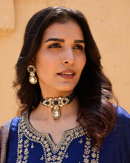 Woman wearing traditional jewelry and a blue embroidered outfit against a beige wall.