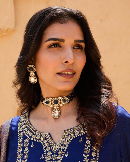 Woman wearing traditional jewelry and a blue embroidered outfit against a beige wall.